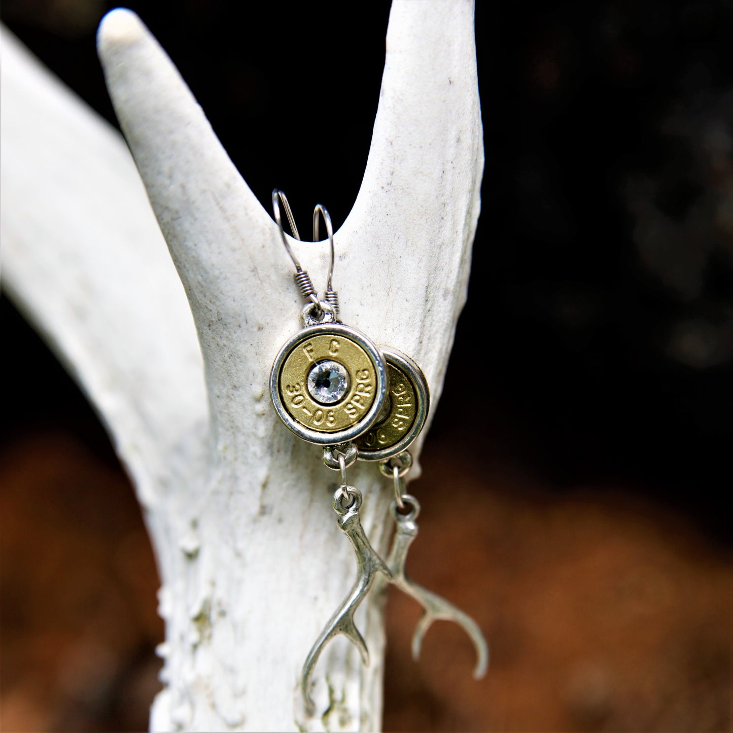 A pair of silver-plated pewter earrings featuring a rifle caliber headstamp and a sparkling Swarovski crystal, displayed on a white antler.